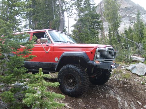 1989 Ford Bronco - A red Jeep Wagoneer navigates a challenging off-road path through a forest with a mountain in the background.