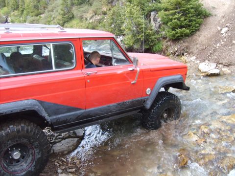 1989 Ford Bronco - A red Jeep Cherokee XJ navigates a rocky stream bed with a driver inside.