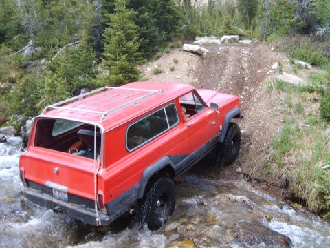 1989 Ford Bronco - This red SUV navigates a rocky stream bed on an off-road trail surrounded by trees.