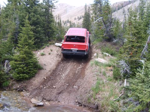 1989 Ford Bronco - Red SUV navigates a challenging dirt path through a wooded mountain area.