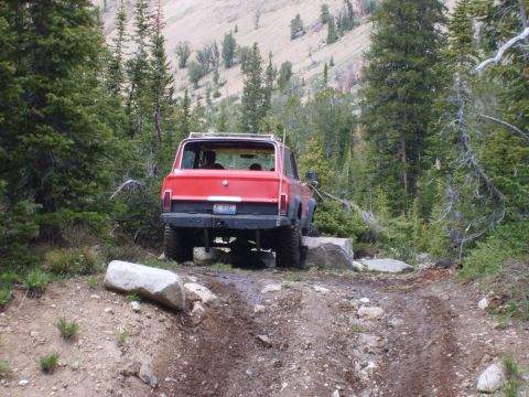 1989 Ford Bronco - A red SUV drives over a large rock on a muddy, off-road trail surrounded by trees.