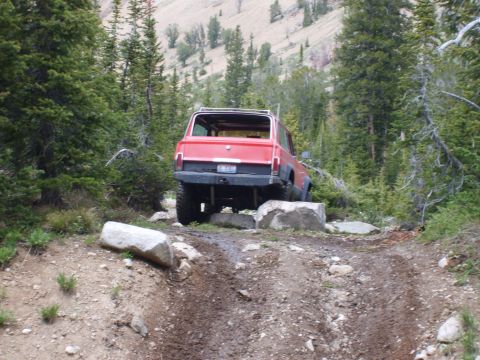 1989 Ford Bronco - A red SUV is shown from the rear, driving over rocks on a muddy trail surrounded by trees.