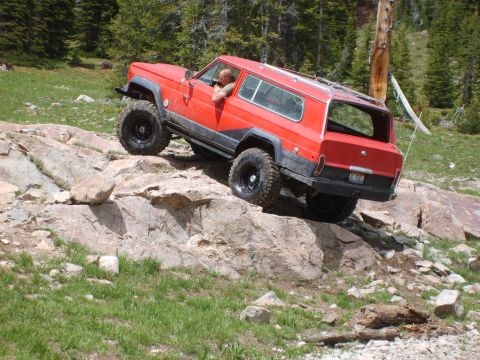 1989 Ford Bronco - Red Jeep Cherokee Chief navigates a rocky trail with a driver visible.