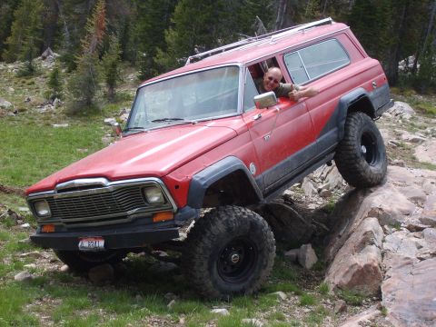 1989 Ford Bronco - This red Jeep Wagoneer is tackling a challenging rocky trail with its driver looking on.