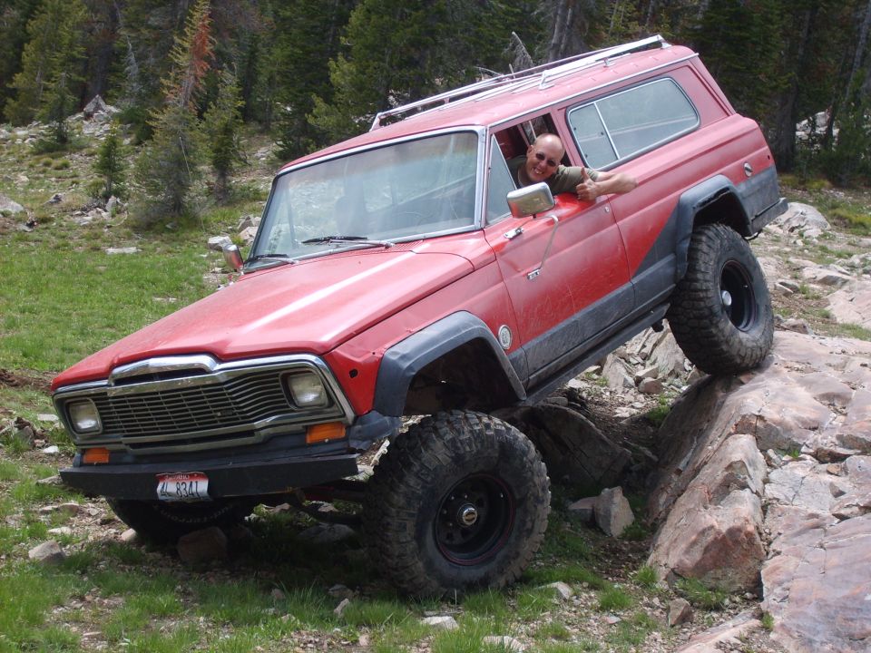 Red Jeep Wagoneer with large tires navigating rocky terrain. Driver is visible through the window