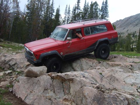 1989 Ford Bronco - A red Jeep Cherokee Chief navigates a rocky trail in a mountainous, wooded area.