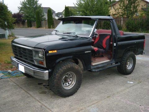 1985 Ford Bronco - This black SUV features a removable top and an open door revealing its red and black interior seats.