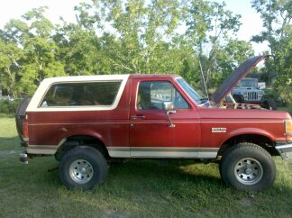 1988 Ford Bronco - A red Ford Bronco with its hood up sits in a grassy area with trees in the background. Another vehicle is parked behind it.