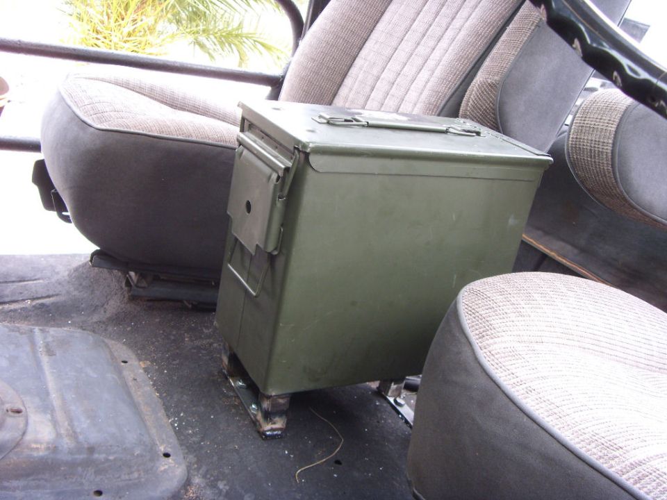 Olive ammo can bolted between gray cloth seats on bare floor of a truck cab