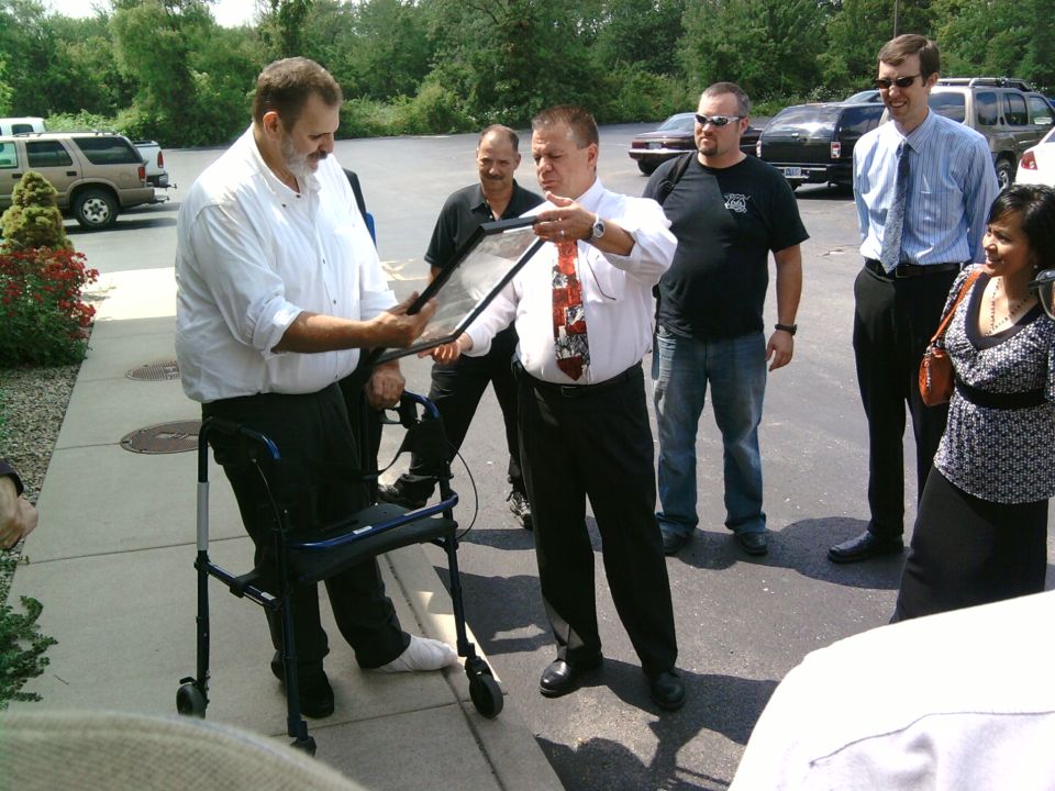 Man in white shirt hands a framed object to another man, with several people watching