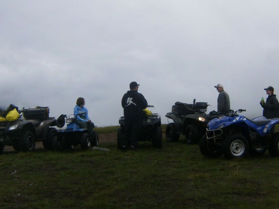 Several ATVs parked on a grassy hill with people standing and sitting around them under a cloudy sky