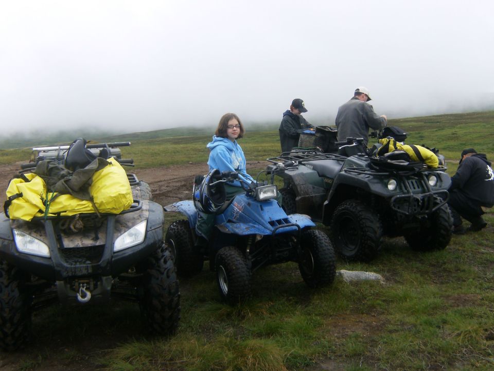 Several ATVs parked on a muddy field with people around. One ATV has yellow bags strapped to the front