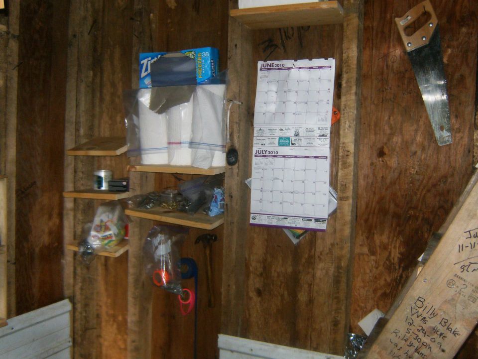 Interior of a wooden shed with shelves holding paper towels, a saw, and a calendar showing June and July 2010