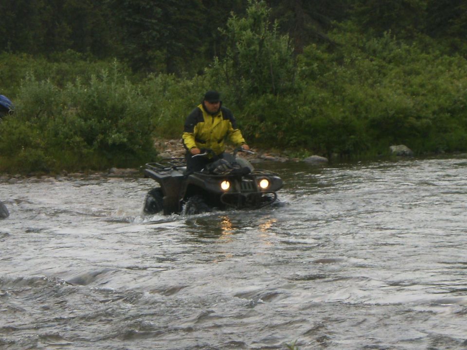 All-terrain vehicle with rider crossing a shallow river, front view. Water splashes around the vehicle