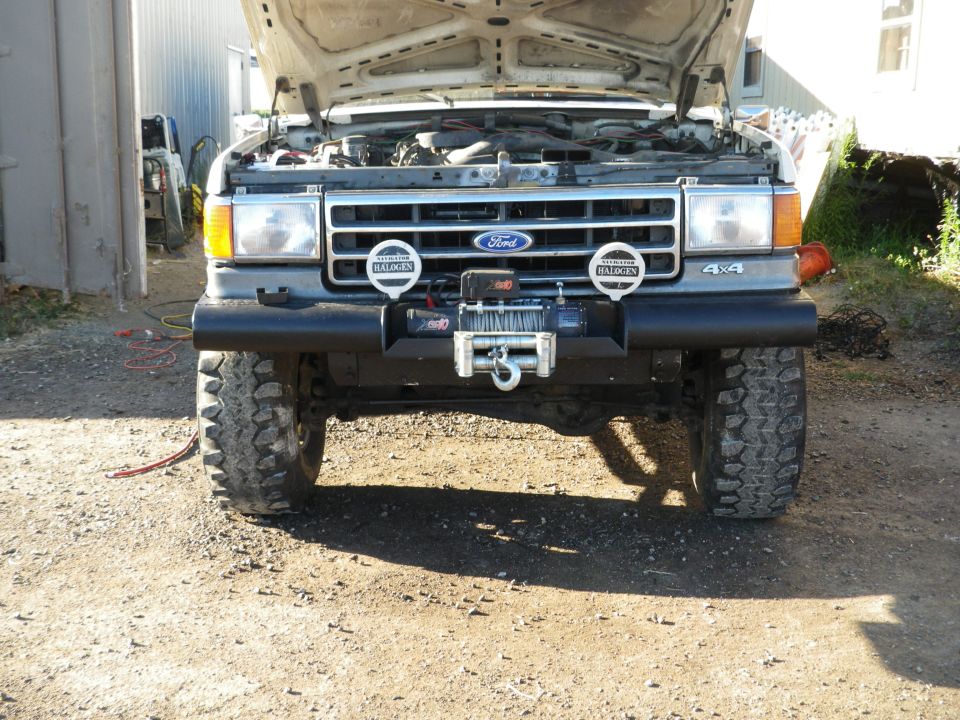 Front view of a Ford Bronco with hood open, showing engine and a winch mounted on the bumper. It has large off-road tires