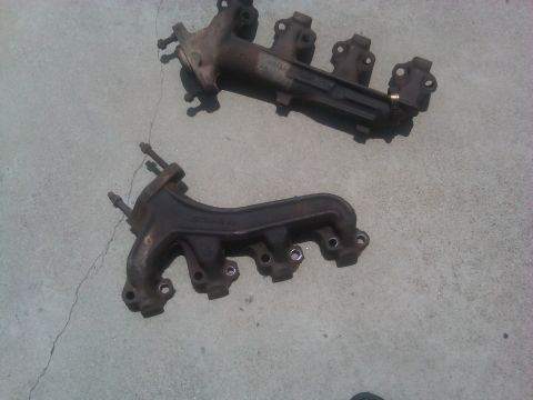 1986 Ford Bronco - Two exhaust manifolds are shown resting on a concrete surface. One is darker and appears older.