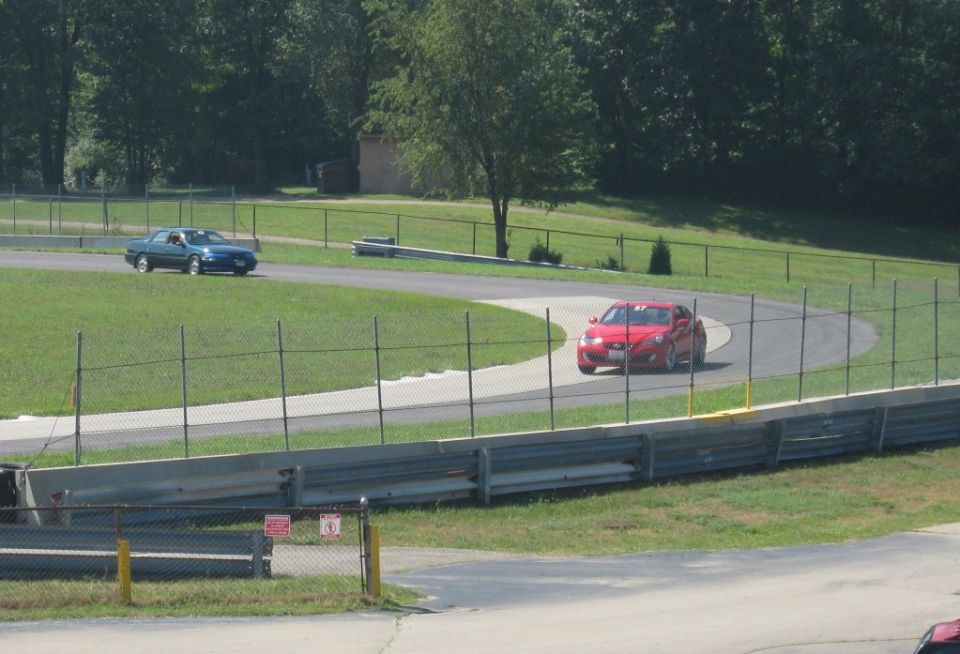 Red coupe rounding a track curve behind fence and guardrail, blue sedan in background
