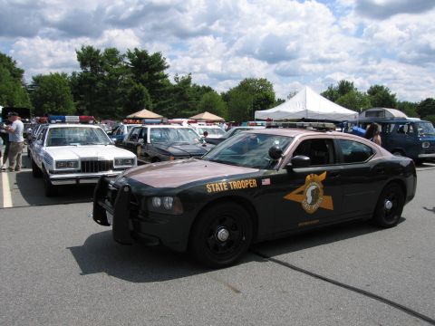 1991 Ford LTD Crown Victoria - dark green Dodge Charger state trooper cruiser with push bar and gold emblem parked among other police cars at an outdoor show.