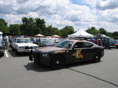 1991 Ford LTD Crown Victoria - Front three-quarter view of a dark green Dodge Charger marked State Trooper, lined up with vintage police cars under a cloudy sky.
