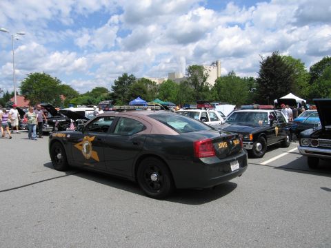 1991 Ford LTD Crown Victoria - Rear quarter view of a dark green Dodge Charger state police cruiser beside a black sedan in a busy outdoor car show lot.