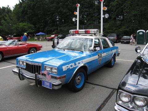 1991 Ford LTD Crown Victoria - blue and white Dodge police sedan marked POLICE with tall roof lights and brush guard, parked at an outdoor car show.