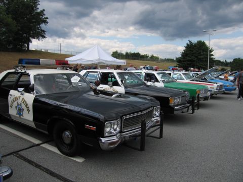1991 Ford LTD Crown Victoria - black and white highway patrol cruiser heads a long row of vintage police cars parked at an outdoor show under cloudy skies.