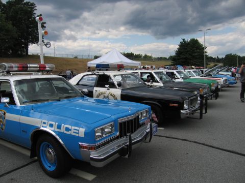 1991 Ford LTD Crown Victoria - blue and white Dodge police car with brush guard leads a lineup of vintage cruisers parked outdoors at a car show.
