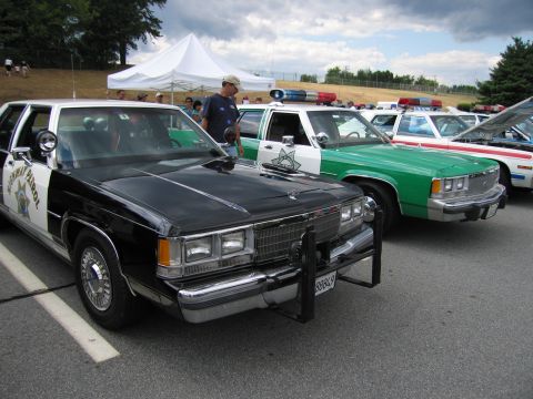 1991 Ford LTD Crown Victoria - black and white Ford highway patrol sedan with push bar parked beside a green and white cruiser at an outdoor car show.