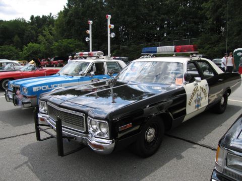 1991 Ford LTD Crown Victoria - black and white Dodge highway patrol car with brush guard parked in front of a blue police sedan at an outdoor car show.