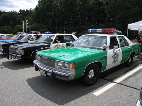 1991 Ford LTD Crown Victoria - green and white Ford sheriff sedan with roof light bar parked beside a black cruiser in a row of police cars at a show.