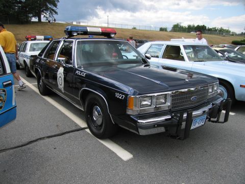 1991 Ford LTD Crown Victoria - dark blue Ford police sedan numbered 1027 with a brush guard and roof light bar parked among cruisers at an outdoor show.