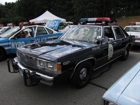 1991 Ford LTD Crown Victoria - dark blue Ford police car numbered 1027 with brush guard parked beside a blue NYPD-marked cruiser at an outdoor show.