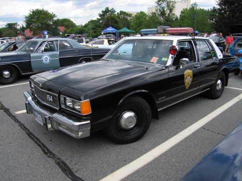 1991 Ford LTD Crown Victoria - black-and-white vintage police sedan with a roof light bar and a gold star door shield sits at a public-safety vehicle show.