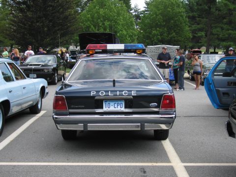 1991 Ford LTD Crown Victoria - rear of a dark police sedan reads POLICE with a roof light bar and license plate 10-CPD, surrounded by spectators on grass.