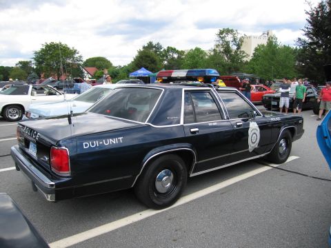 1991 Ford LTD Crown Victoria - dark four-door police sedan marked POLICE and DUI-UNIT with a red-blue roof light bar sits among show cars and onlookers.