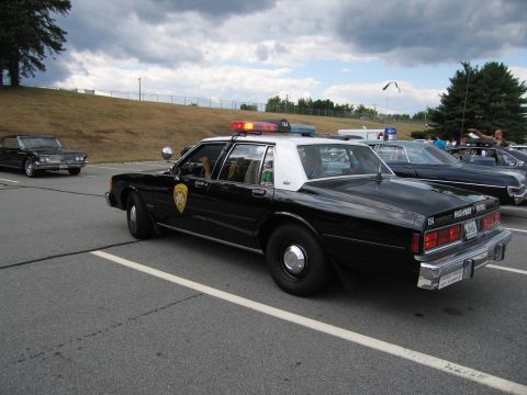 1991 Ford LTD Crown Victoria - black-and-white police sedan with a red roof light bar and gold star shield is parked on a hillside lot among other vehicles.