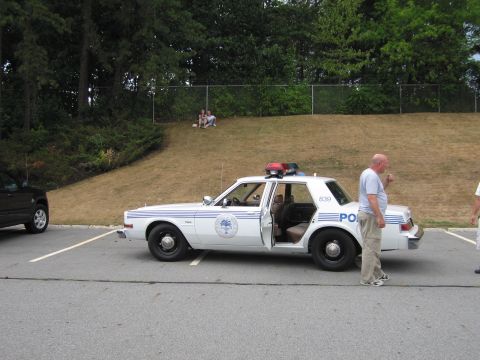 1991 Ford LTD Crown Victoria - white marked police sedan with a palmetto-tree door emblem and roof light bar sits in a lot as a man stands beside it.
