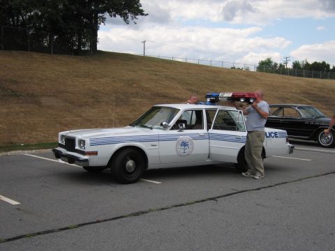 1991 Ford LTD Crown Victoria - white police sedan with blue stripes and a round palmetto door emblem sits as two men handle a roof light bar.