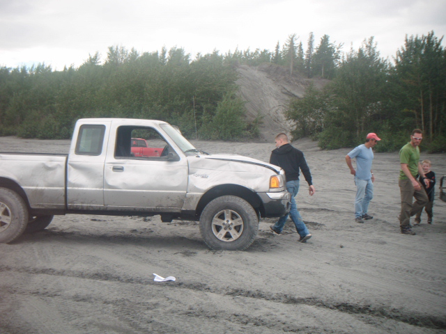 Dented silver extended-cab compact pickup with people in a gravel pit