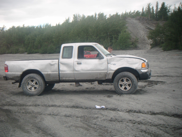 Silver pickup truck with damage to the front fender and hood. A red vehicle is visible in the passenger side window