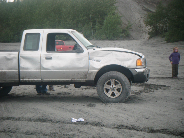 Muddy white extended-cab pickup with dented front fender parked in a sandy gravel pit