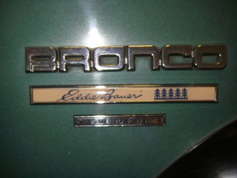 1990 Ford Bronco - Close-up of chrome 'BRONCO' emblem on a dark green metallic body panel, with an 'Eddie Bauer' script badge featuring pine tree graphics and…