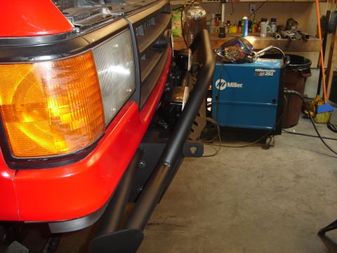 1989 Ford Bronco - Close-up of a red Ford Bronco's front end, showing the grille and aftermarket bumper. A Miller welding machine is visible in the background.