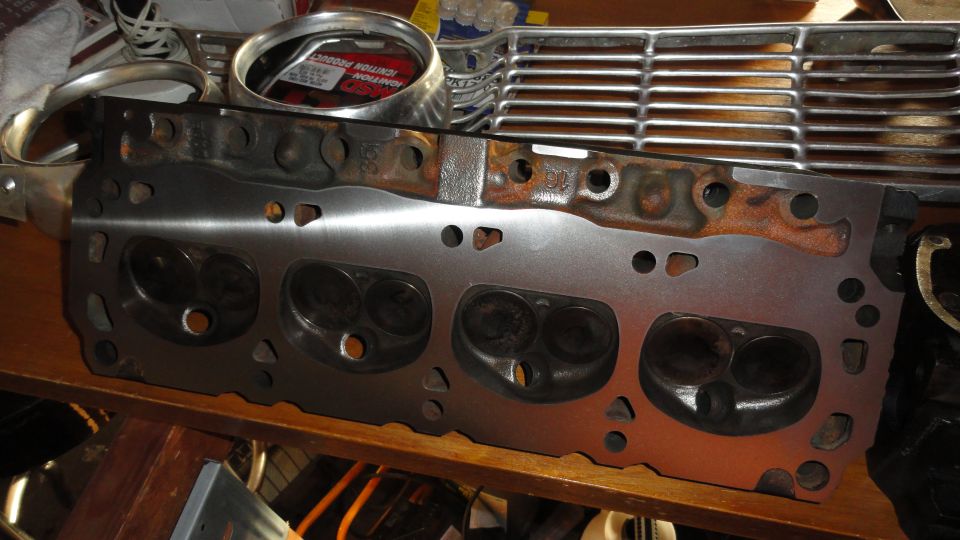 Engine cylinder head with four intake valves visible, resting on a wooden surface next to a chrome grille