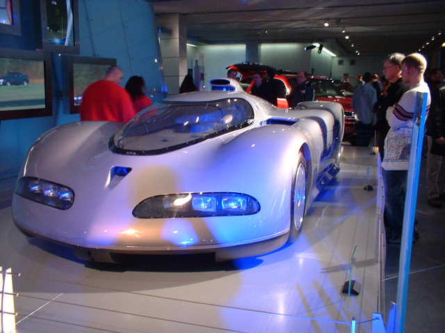 Silver concept car with blue headlights on display, people watching