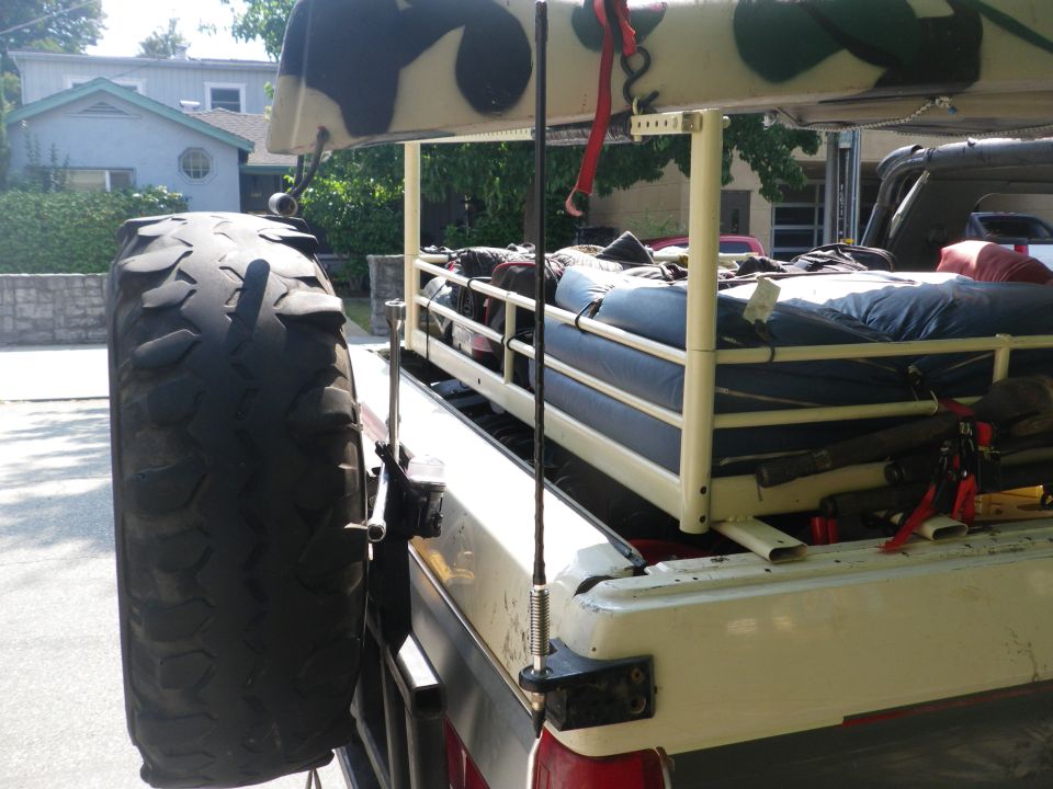 Pickup truck bed loaded with camping gear, spare tire mounted on the side. Another vehicle is visible in the background