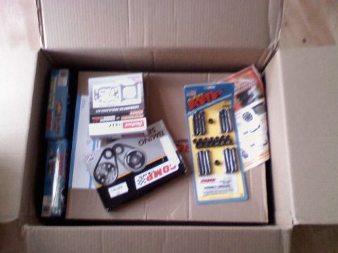 1979 Ford Bronco - An open cardboard shipping box on a wood floor filled with newly delivered engine build parts, including a boxed ARP head/fastener kit in b…