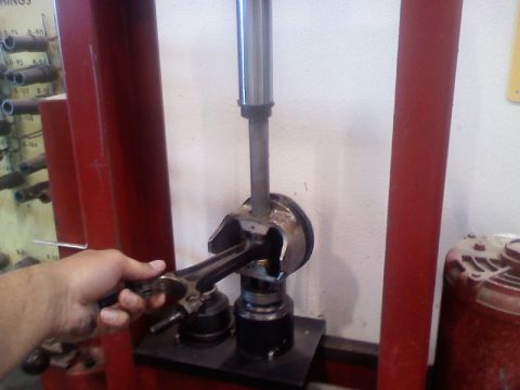 1979 Ford Bronco - A hand steadies a piston and connecting rod assembly under the ram of a red hydraulic shop press, with the piston skirt clamped in a fixtur…