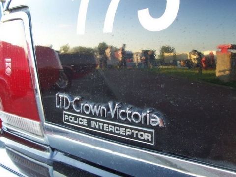 1991 Ford Crown Victoria - Rear badge of a black 1991 Ford Crown Victoria Police Interceptor, reflecting a crowd and vehicles.