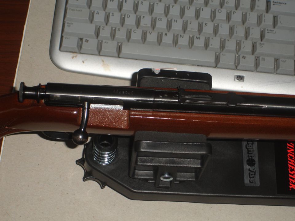 Close-up of a rifle with wooden stock on a black stand, keyboard in background. Text 'SUPERMOTORS.NET' visible